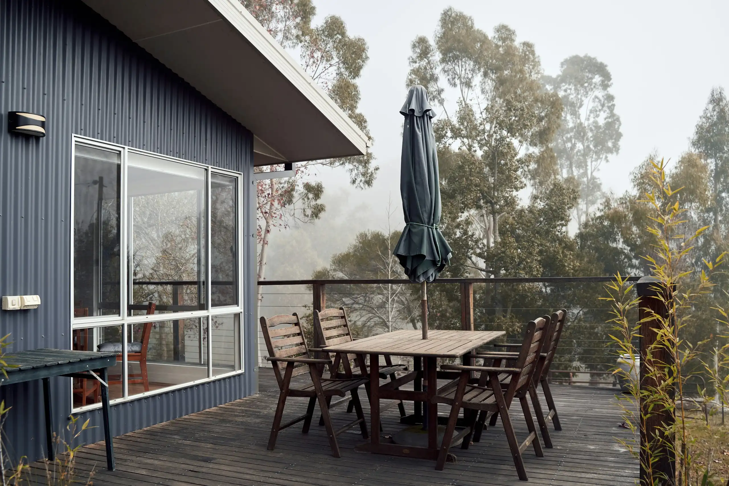 A timber deck with an outdoor table beside a house clad in dark metal sheeting, gum trees behind
