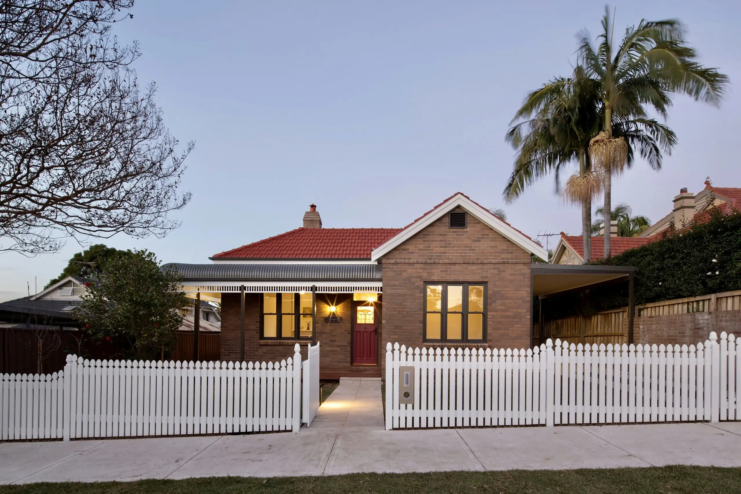 A red-brick bungalow at dusk behind a freshly painted white picket fence, its windows lit and a palm beside it