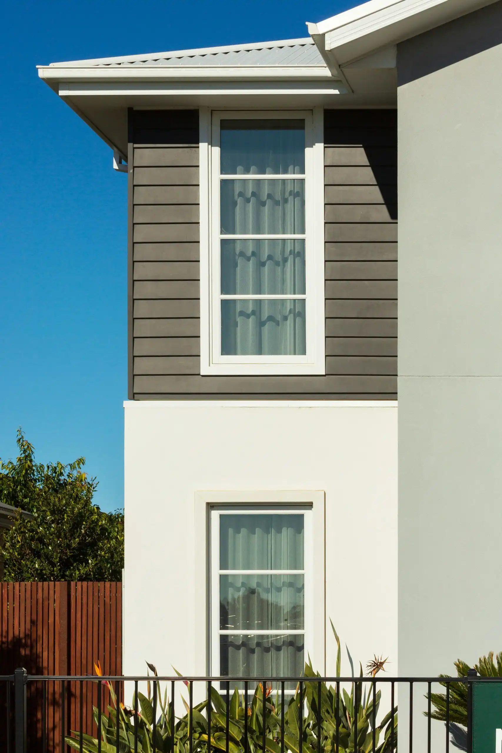 A two-storey house with dark grey weatherboards over white render and white window trims
